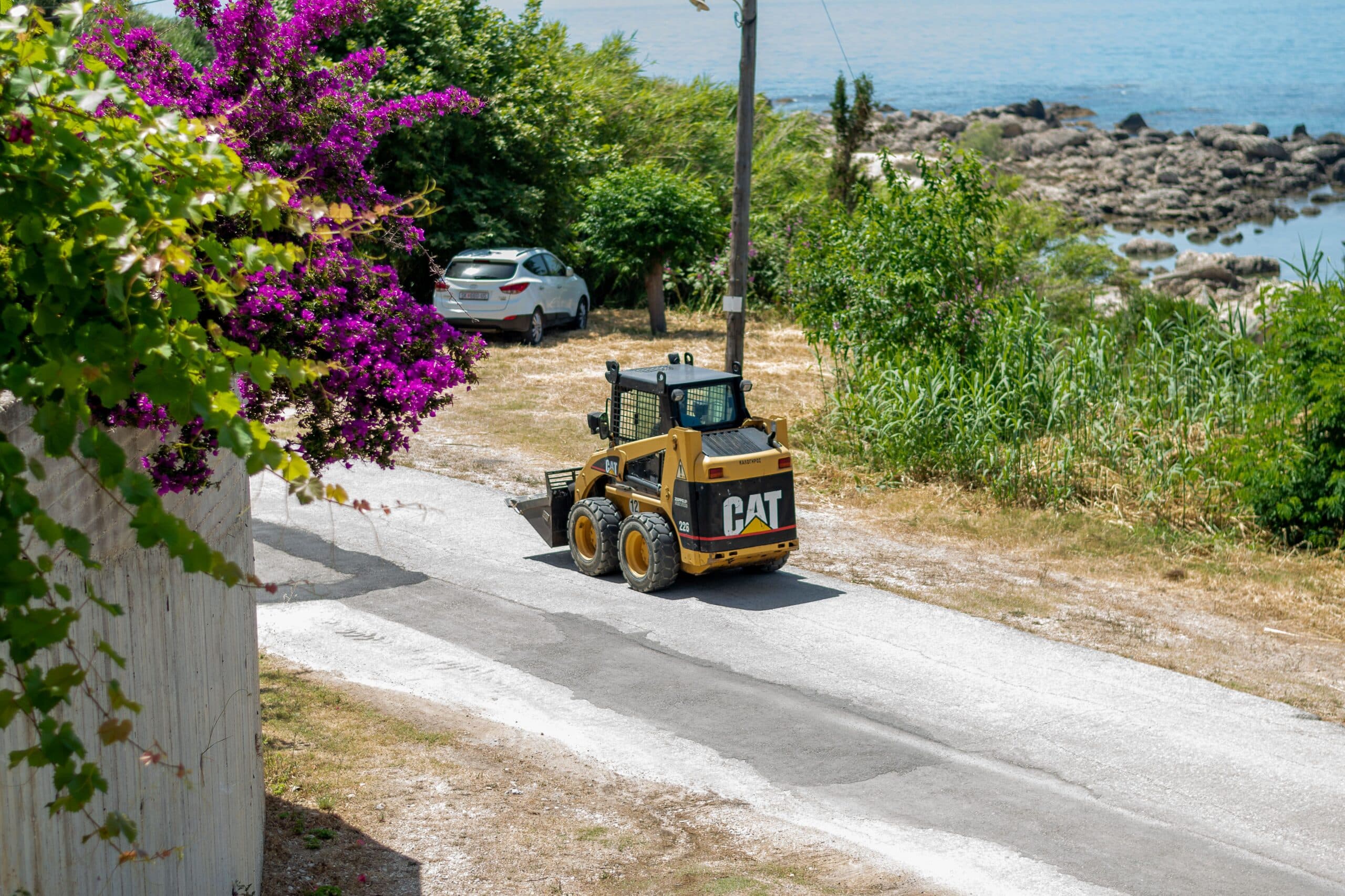 A compact skid steer loader navigating a rural roadside verge next to dense vegetation and trees